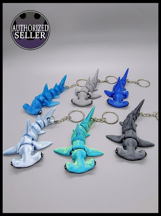 Assorted 3D printed baby hammerhead shark keychains in blue, green, gray, and marble colors – articulated fidget toy design with keychains.