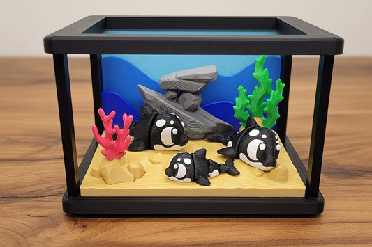 A black rectangular display case containing a miniature underwater scene with three black-and-white orca toys, gray rocks, green seaweed, and pink coral on a sandy base.