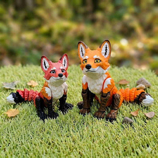Two 3D printed articulated fox figurines sitting on green grass — a smaller red fox with cartoon eyes on the left and a larger orange fox with gold eyes on the right, both with multicolor segmented tails
