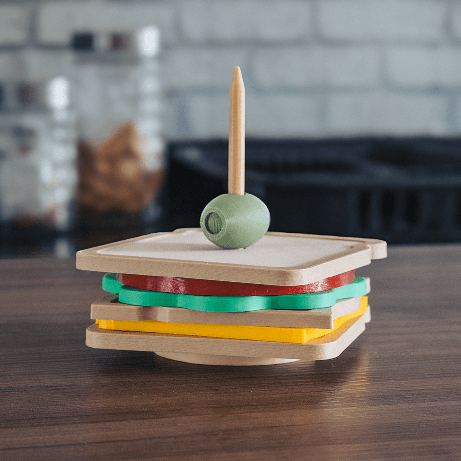 3D printed club sandwich coaster set stacked on a dark wood surface, showing layered square coasters in tan, red, green, and yellow with a decorative green olive and toothpick on top, kitchen background