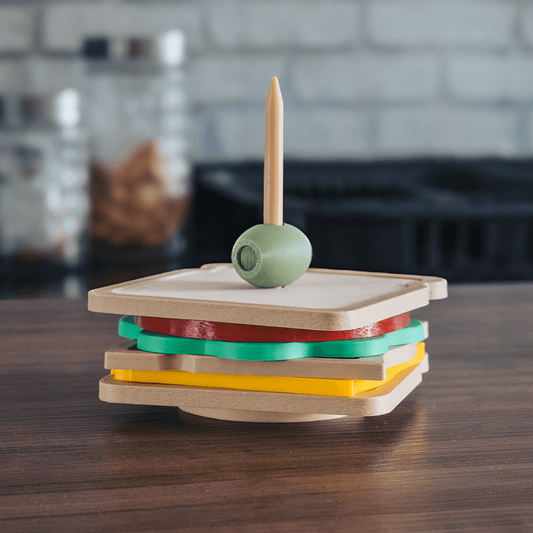 3D printed club sandwich coaster set stacked on a dark wood surface, showing layered square coasters in tan, red, green, and yellow with a decorative green olive and toothpick on top, kitchen background