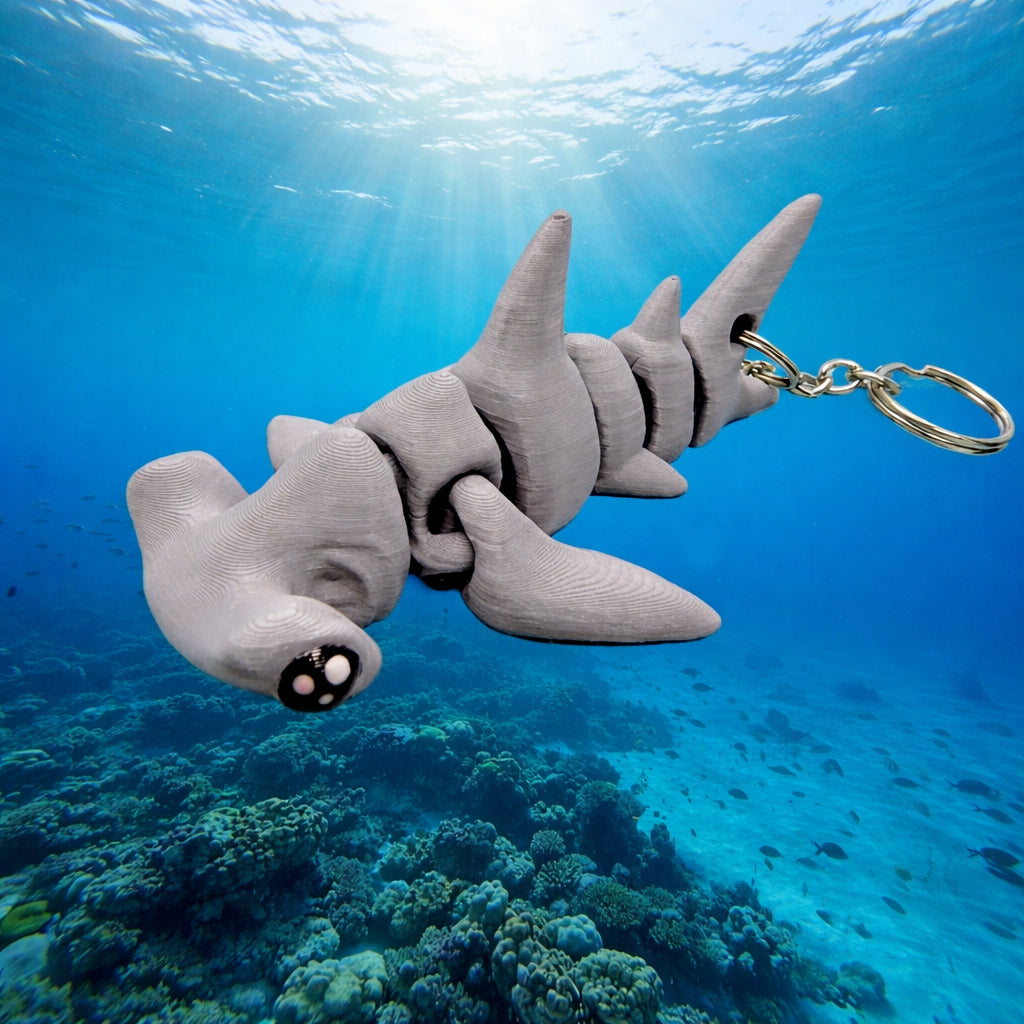 Gray articulated baby hammerhead shark toy with segmented flexible body and fins, shown underwater against a bright blue ocean background with sun rays filtering through the water and a coral reef below. The shark has black eyes with white highlights and a silver metal keychain attached to the tail.