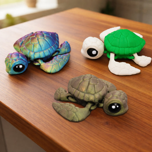 Three articulated baby sea turtle toys displayed on a brown surface, shown in rainbow multicolor, green and white, and earthy brown finishes with large black eyes and movable flippers
