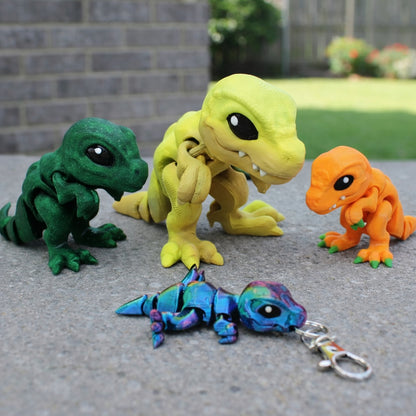 Four articulated baby T Rex toys displayed outdoors on a concrete surface, shown in dark green, yellow, orange, and rainbow multicolor, with one small rainbow T Rex attached to a metal keychain and a blurred brick wall and grassy yard in the background