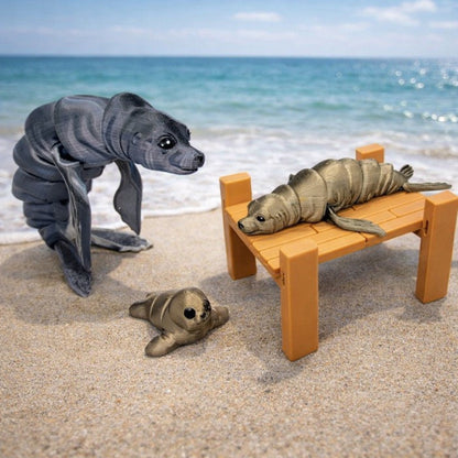 Articulated flexi sea lion toy shown in dark gray, tan, and light brown color variants on a sandy seashore background, with one sea lion resting on an optional dock. Posable 3D printed fidget sea lion available in two sizes with customizable colors.