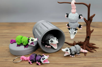 Articulated mini possum toys in grey and a mini possum keychain in purple and black, shown posed on a tabletop with a possum hanging from a tree prop and one peeking from inside a toy trash can, cute sensory fidget figures