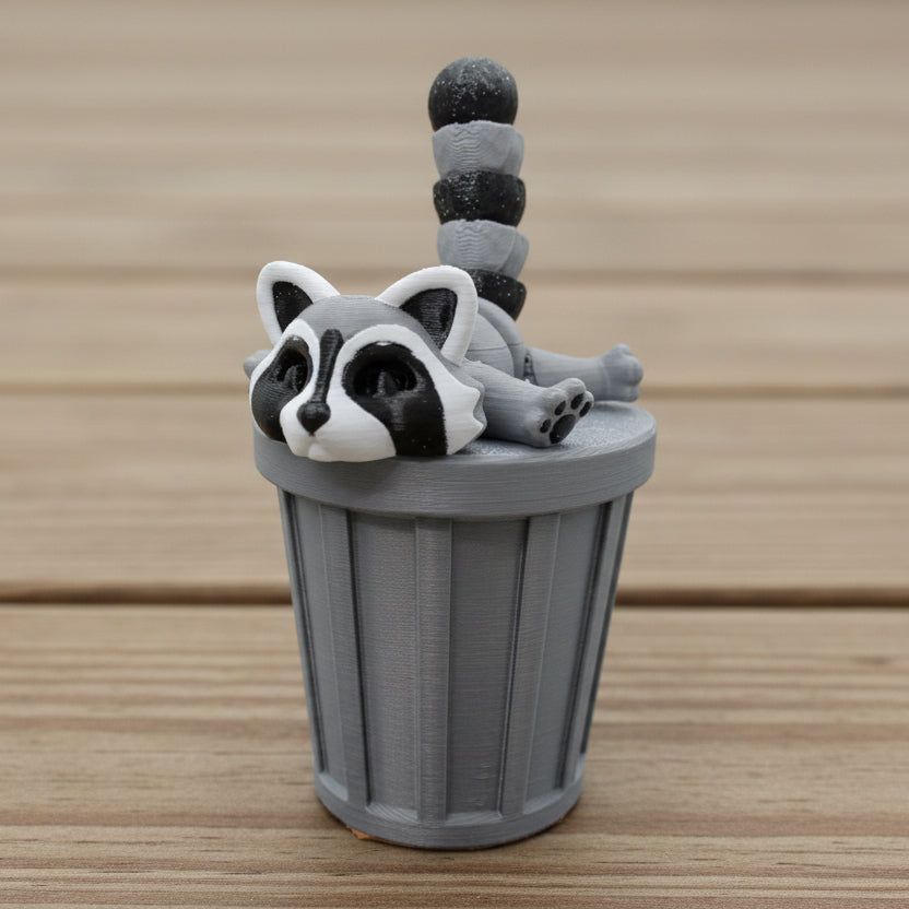 Articulated raccoon fidget toy in light gray and dark gray with black facial markings, posed inside a matching gray trash can accessory on a wooden tabletop