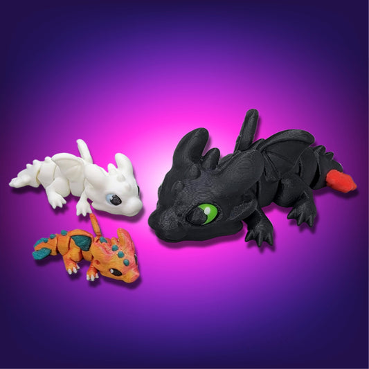 Three small articulated baby dragon figures in different colors, including a black dragon with green eyes and two smaller dragons, displayed on a purple gradient background