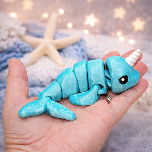 Small articulated blue narwhal toy with a white horn resting in a person’s hand, shown against a soft ocean themed background with a starfish