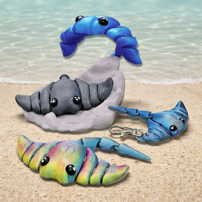 Baby stingray toy or keychain featuring articulated 3D printed stingray figures in blue gray and rainbow colors displayed on a sandy beach background including a keychain attachment and flexible segments for sensory play gifts or accessories