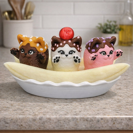 Three banana split kitty clicker toys displayed in a white banana bowl on a kitchen countertop. The cats feature chocolate brown, cream white, and pastel pink bodies with sprinkle details, a red cherry topper, and raised paws. The banana and bowl are fully customizable colors. Nine different kitty color options available.