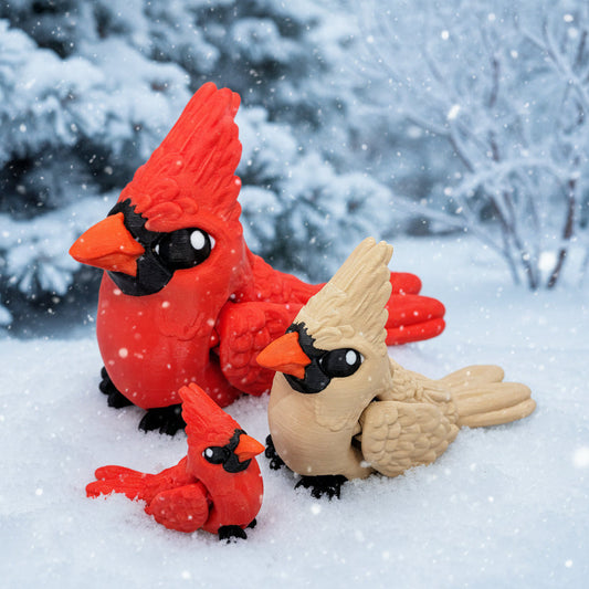 Three cardinal bird toys in red and tan colors resting in snow, including two larger adult birds and one smaller baby bird against a winter forest background