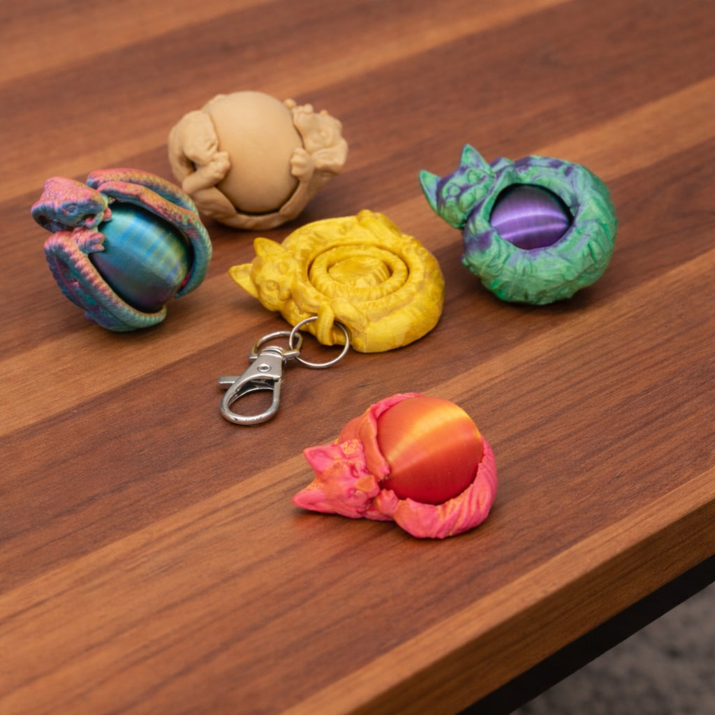 Five creature themed fidget spinners displayed on a wooden surface, including dragon styled figures in multicolor, beige, green and purple, yellow with a metal keychain clip, and red and orange, each curled around a central spinning core