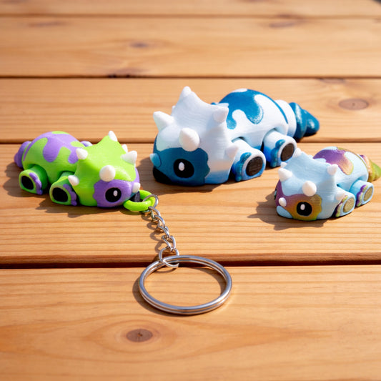 Three custom baby triceratops dino toys with articulated bodies, shown in green and purple, blue and white, and rainbow colors, arranged on a brown surface with one attached to a metal keychain ring