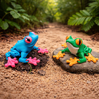 Two articulated frog toys in custom colors, one blue with pink feet and one green with orange feet, posed on rocks against a blurred natural forest background.