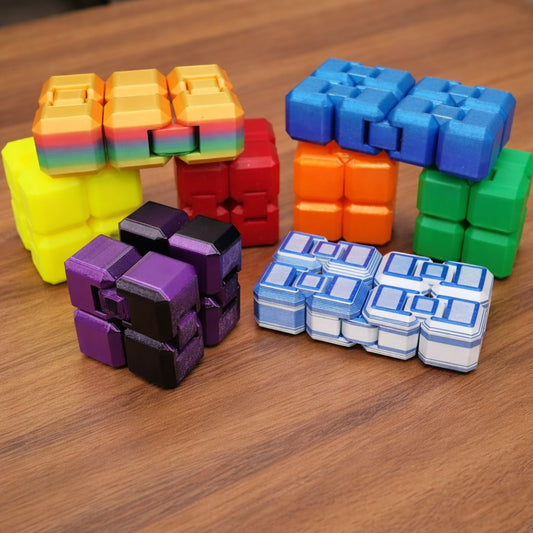 Multiple custom infinity fidget cubes in assorted colors, including rainbow, blue, orange, green, purple, black, and white, arranged on a brown surface in different folded positions
