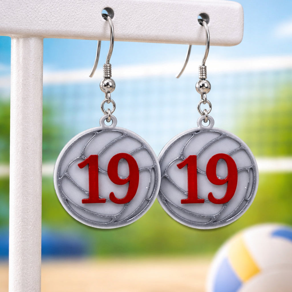 Pair of earrings with '19' design on a stand with a blurred beach background