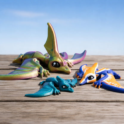 Articulated flappy pterodactyl and nyctosaurus toys in colorful finishes displayed on a wooden surface under a blue sky.