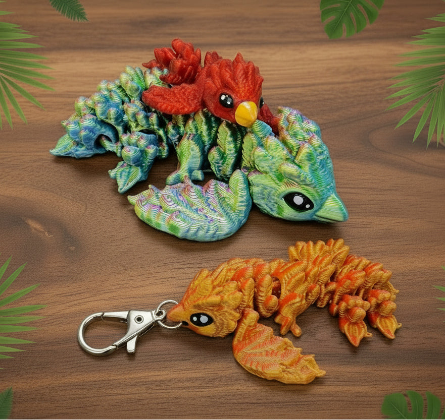 Two floppy baby phoenix figures with articulated bodies displayed on a brown surface, including a larger multicolor green and red phoenix toy and a smaller orange phoenix keychain attached to a silver metal clasp