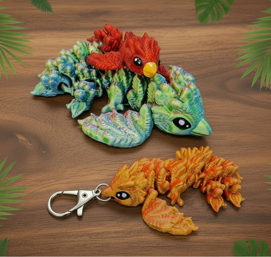 Two floppy baby phoenix figures with articulated bodies displayed on a brown surface, including a larger multicolor green and red phoenix toy and a smaller orange phoenix keychain attached to a silver metal clasp