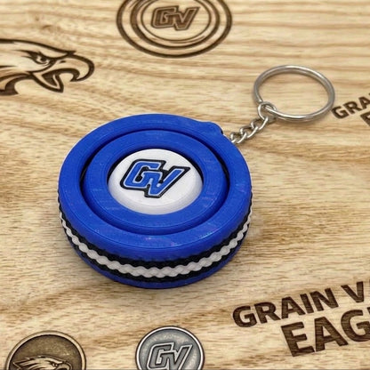 Close-up of a blue-and-white circular fidget spinner keychain with a blue GV logo on a wooden Grain Valley Eagles sign.