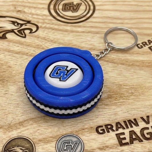 Close-up of a blue-and-white circular fidget spinner keychain with a blue GV logo on a wooden Grain Valley Eagles sign.