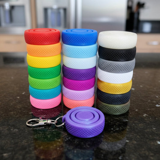 Stacks of round gyroid fidget spinners in multiple colors arranged on a dark countertop, including red, orange, yellow, green, blue, purple, white, gray, and black, with one purple gyroid spinner attached to a silver metal keychain clip in the foreground
