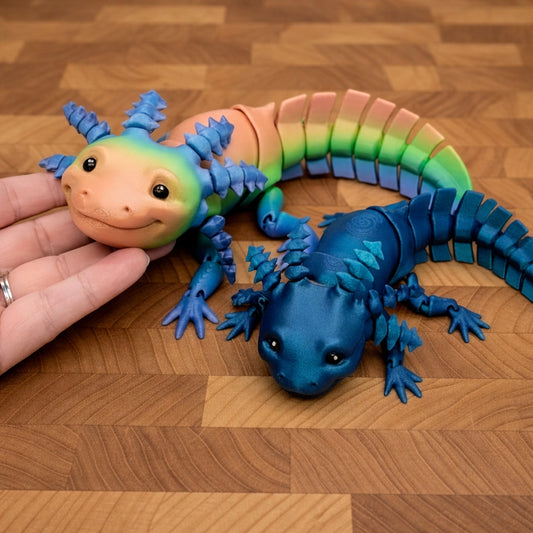 Two articulated axolotl fidget toys displayed on a warm wooden tabletop, featuring one pastel rainbow axolotl with peach, green, and pink tones and bright blue frilled gills, alongside a deep blue axolotl with textured scales and matching blue gills