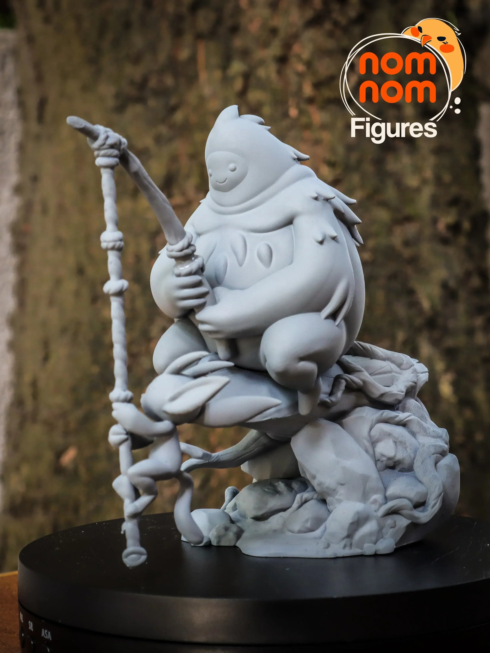 White figurine of a character with a staff on a black base, branded 'nom nom Figures'.