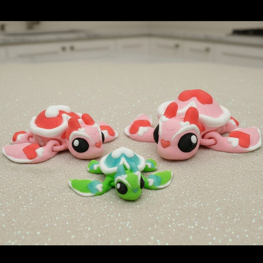 Three small sea turtle figures with heart shaped patterns in pink, red, and green resting on a light glittered surface