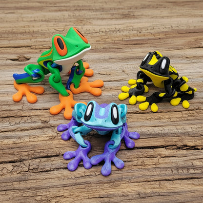 Three colorful frog figurines arranged on a rustic wooden surface: a green frog with orange feet and blue back at upper left, a blue and purple frog in front, and a black and yellow frog at upper right.