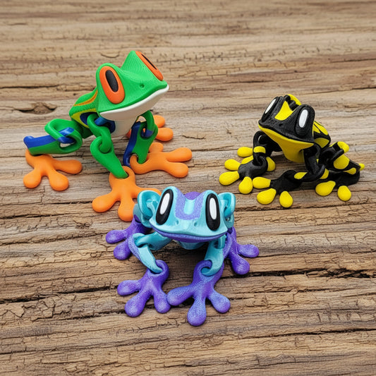 Three colorful frog figurines arranged on a rustic wooden surface: a green frog with orange feet and blue back at upper left, a blue and purple frog in front, and a black and yellow frog at upper right.
