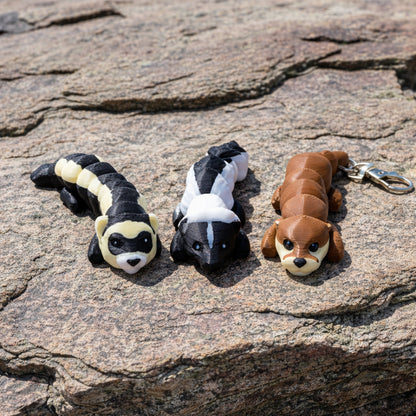 Three mini articulated animal figures displayed on a rock surface outdoors, including a black and cream ferret, a black and white skunk, and a brown otter with a metal keychain clip attached