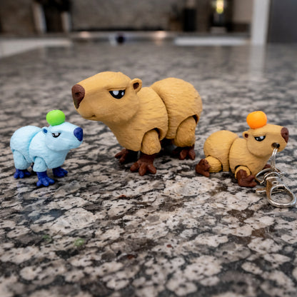 Three mini articulated capybara figures displayed on a gray granite countertop, including a small light blue capybara with a green ball on its head, a larger tan capybara standing in the center, and a tan capybara keychain with an orange ball on its head attached to a silver metal clasp