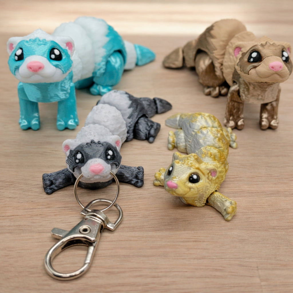 Five mini ferret figures with articulated bodies, shown in teal and white, brown, black and white with a metal keychain clip, and metallic gold, arranged on a neutral surface