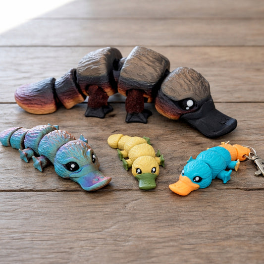 Articulated mini platypus toy figures in multiple colors displayed on a wooden surface, including keychain sized platypus toys and a larger flexi platypus model.