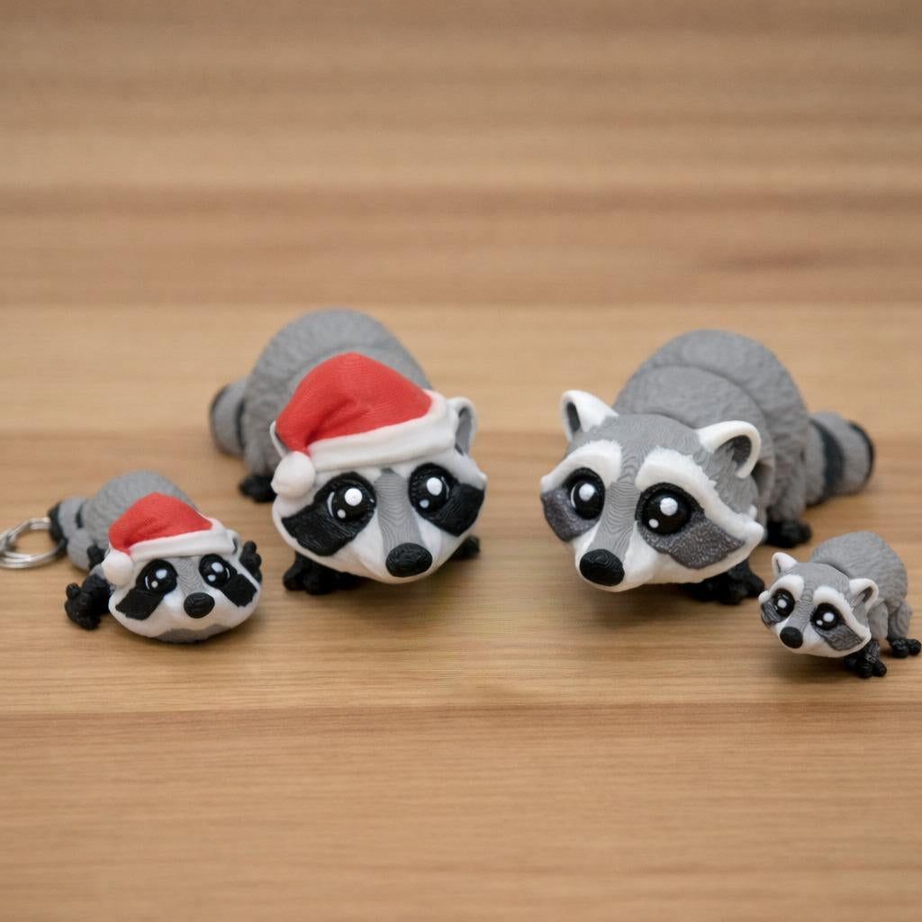 Mini raccoon toy or keychain featuring a small gray and black 3D printed raccoon with big eyes shown in multiple sizes on a brown surface including a keychain version and miniature desk toy for gifts sensory play or accessories