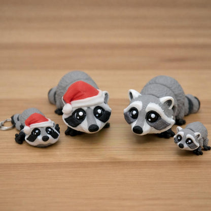 Mini raccoon toy or keychain featuring a small gray and black 3D printed raccoon with big eyes shown in multiple sizes on a brown surface including a keychain version and miniature desk toy for gifts sensory play or accessories