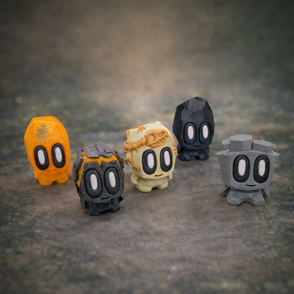 Five small collectible prehistoric rock toys with carved stone textures and expressive eyes arranged on a neutral surface, including an orange, gray, black, and fossil styled figure displayed together as a set.