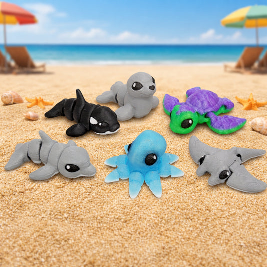 Articulated sea hatchlings toy pack featuring a dolphin, orca, seal, sea turtle, octopus, and hammerhead shark arranged on a sandy beach scene.