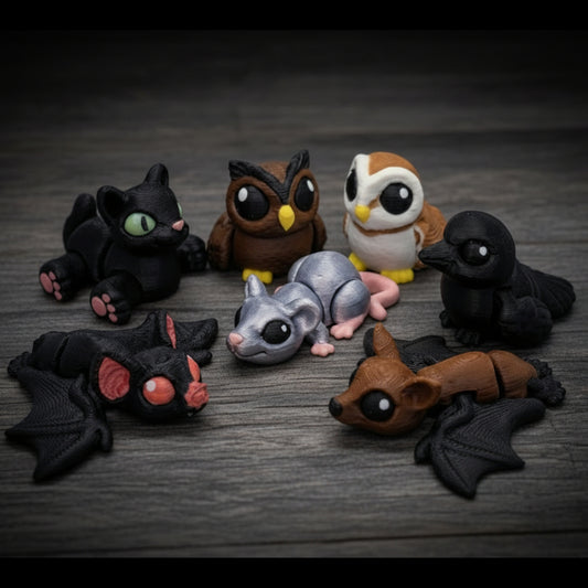 Assorted spooky hatchling animal toys arranged in a circle on a dark wood surface, featuring bats, owls, a mouse, a cat, and other small creatures in black, brown, gray, and white tones.