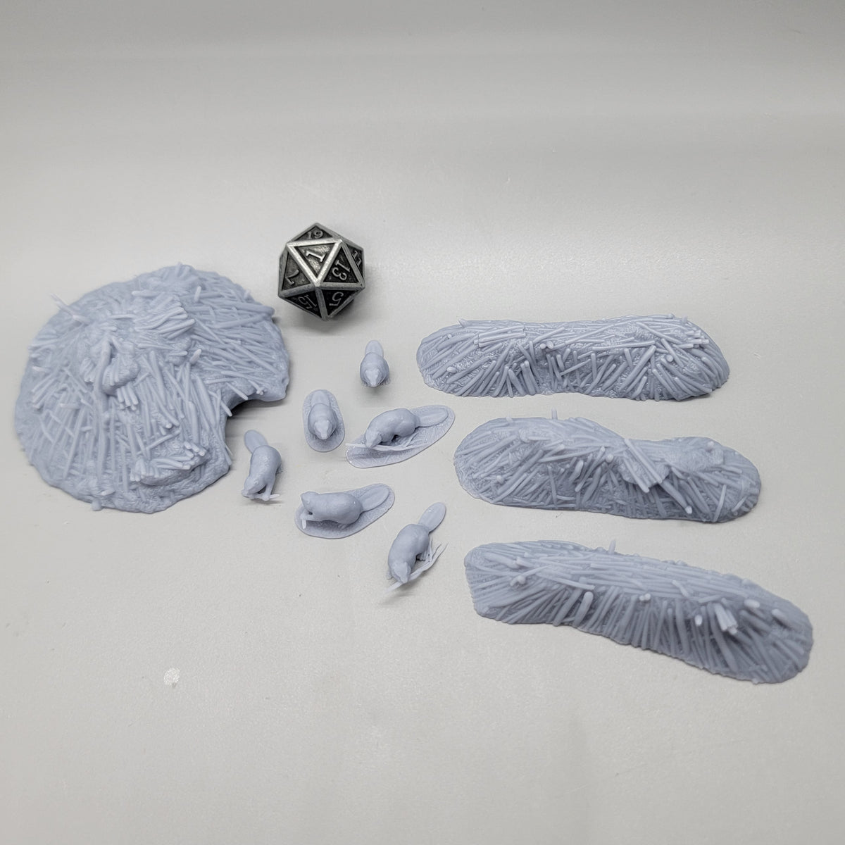 Top-down view of the unpainted resin Beaver Pack scatter set featuring 6 beaver miniatures, 3 dams, 1 lodge, and a D20 die for scale.