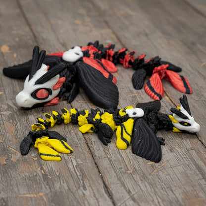 Two 3D printed articulated Death's Head Moth Dragon toys on a weathered wood surface — a larger black and red dragon with white face markings and moth wings on the left, and a smaller black and yellow dragon with white skull markings on the right
