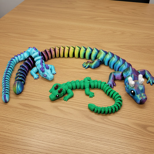 Three 3D printed articulated horned dragon toys displayed on a wood desk — a small blue and purple gradient dragon, a medium solid green dragon with brown horns, and a large blue-green-purple iridescent dragon with white horns