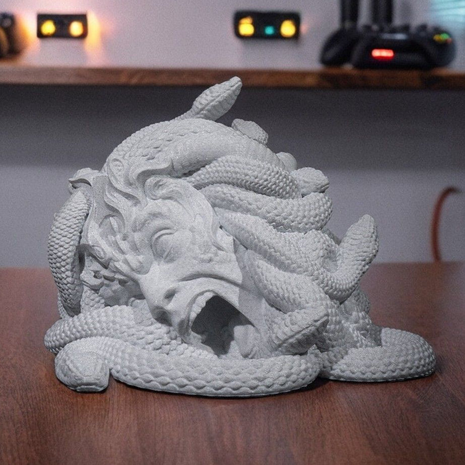 3D printed Medusa dice tower in light grey, featuring a detailed female face with open mouth dice exit surrounded by coiling snakes, displayed on a wood desk with gaming equipment in background