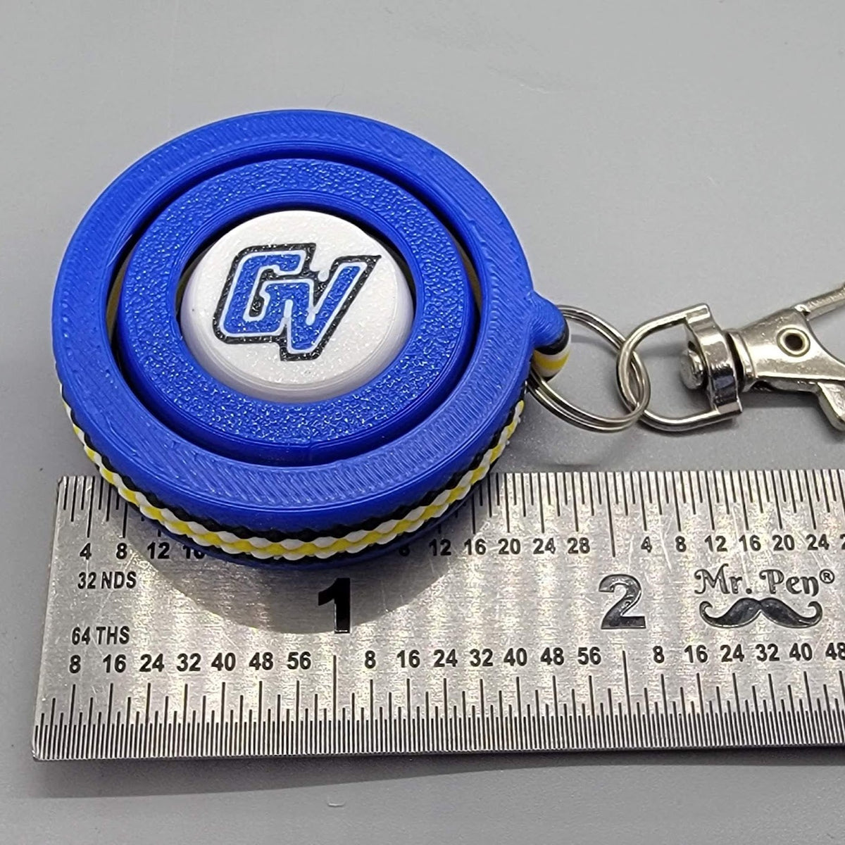 Top-down view of the blue GV 3D printed fidget spinner keychain resting next to a metal ruler showing dimensions