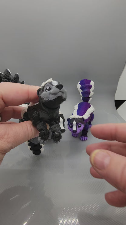 Showcase of 3 small articulated skunk figures, 2 of which are black, grey and white, while the third is white, black, and purple.